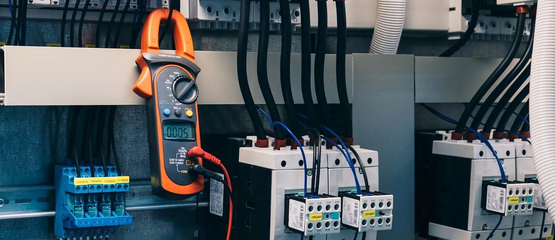 Electrical control panel with wiring, circuit breakers, and a clamp meter hanging inside an open cabinet