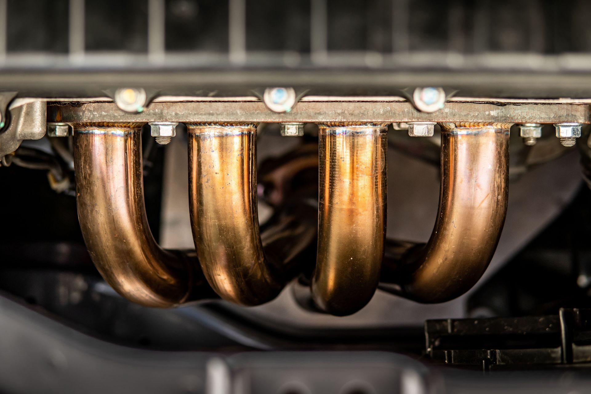 A close-up view of a car engine's four shiny, copper-colored exhaust manifold pipes mounted to the engine block.