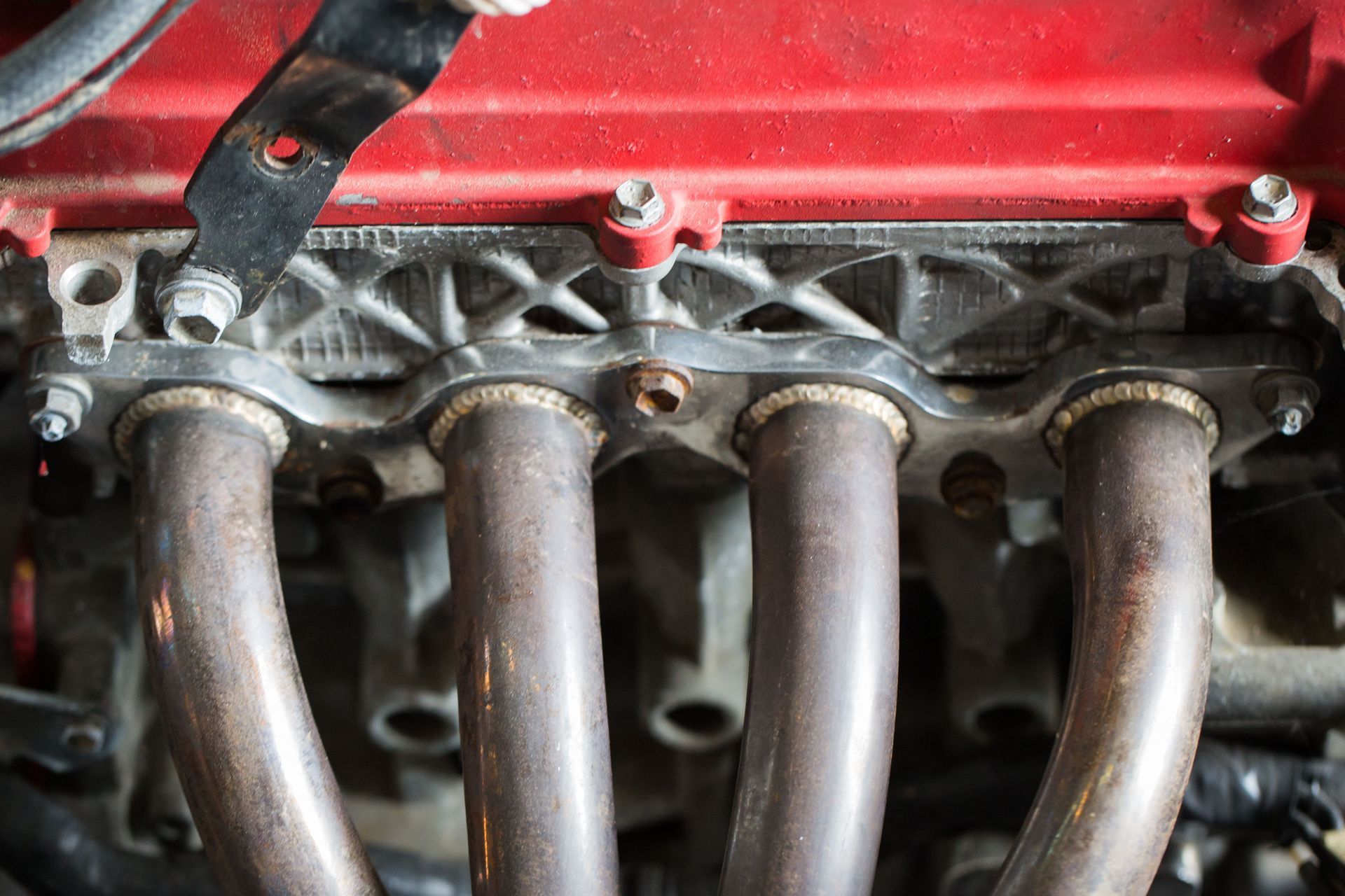 Close-up of a rusty, four-pipe engine exhaust manifold bolted to a red engine block in an automotive engine bay.