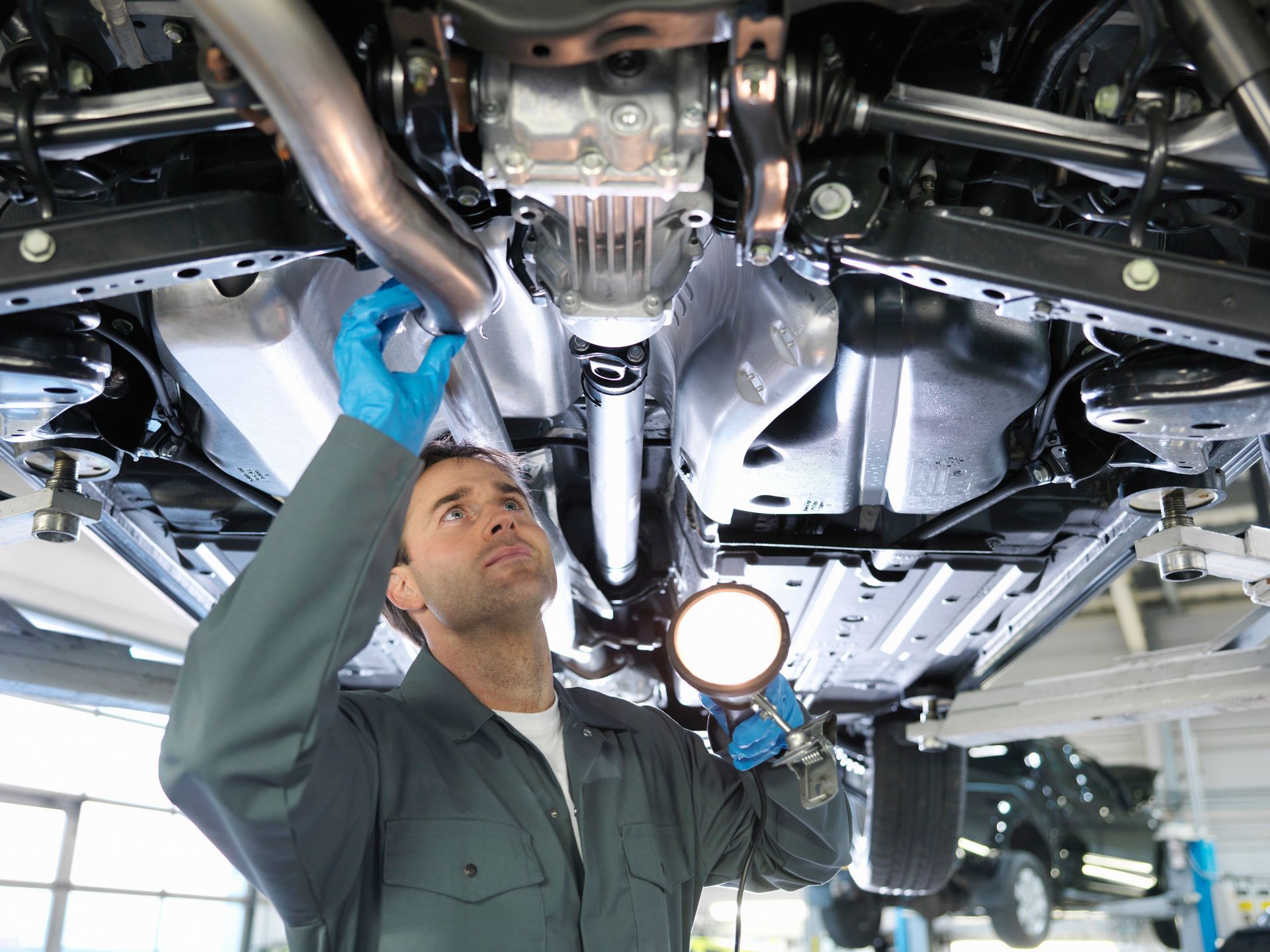 A mechanic in blue gloves inspects the undercarriage of a raised vehicle using a flashlight in a garage.