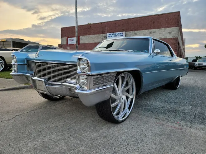 A light blue classic Cadillac parked on a paved lot at sunset, featuring large, modern chrome custom wheels.