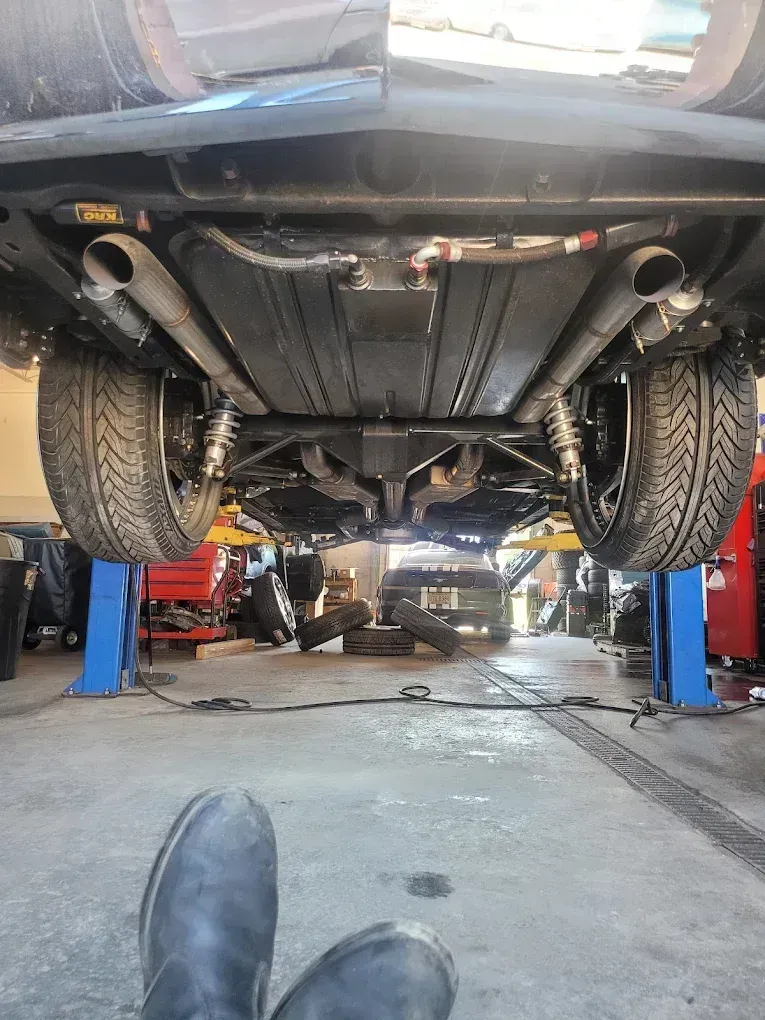 A low-angle view from underneath a car hoisted on a lift, showing the chassis, suspension, tires, and exhaust pipes.