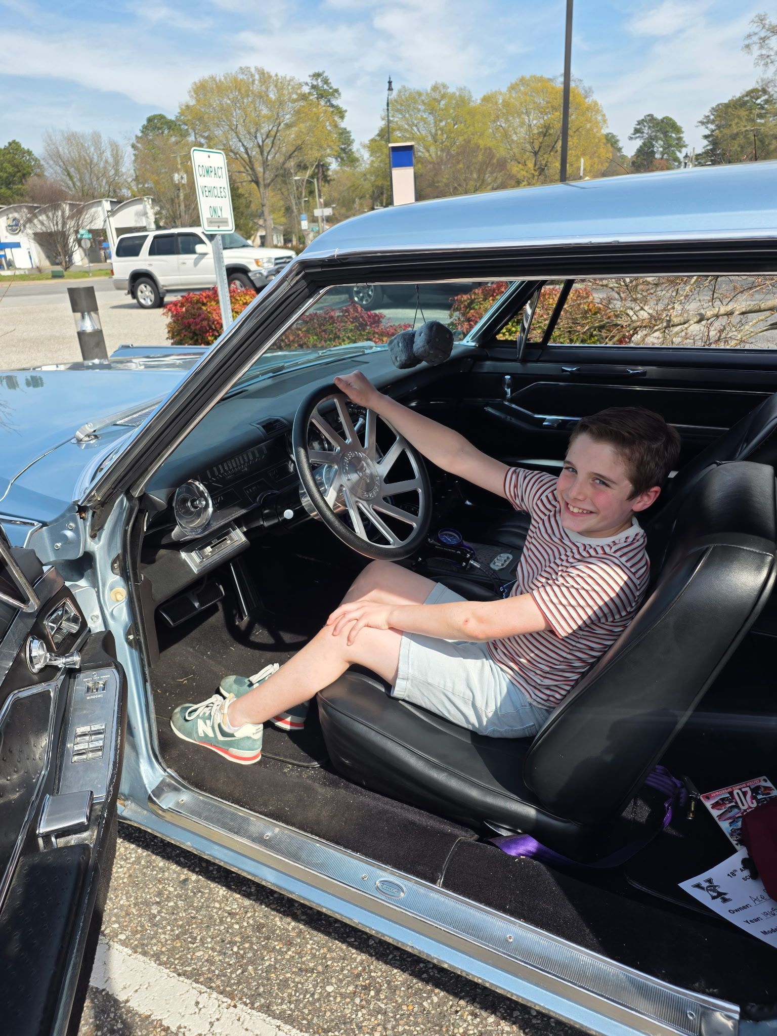 Child sitting in a classic car, smiling and holding the steering wheel at an outdoor event