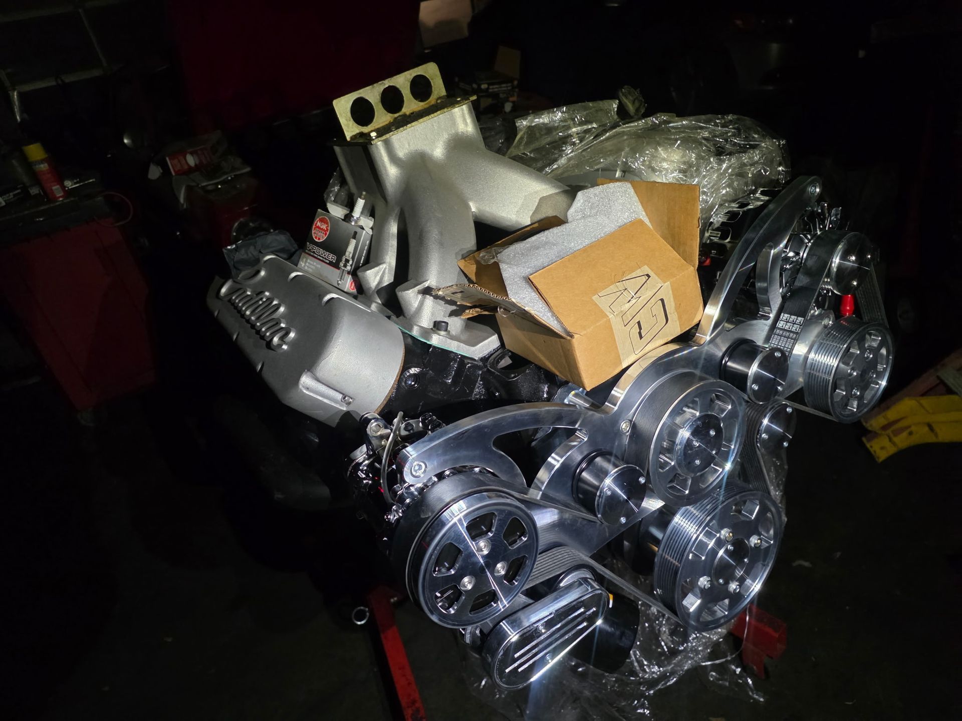 Engine parts and wrapped items piled in a dark storage area, with shiny metal pulleys in front.