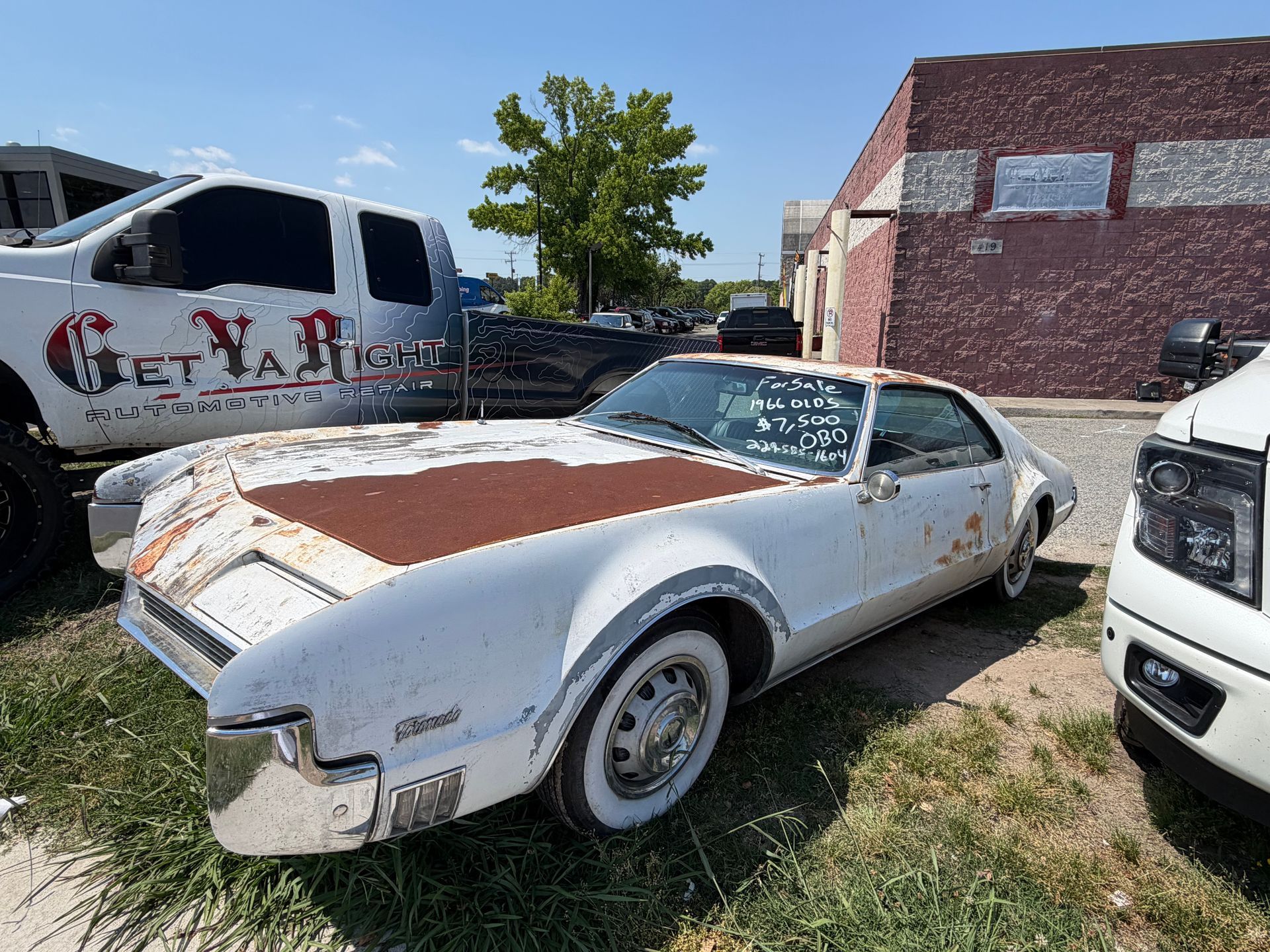 Vintage white-and-brown car parked outdoors beside a work truck and brick building