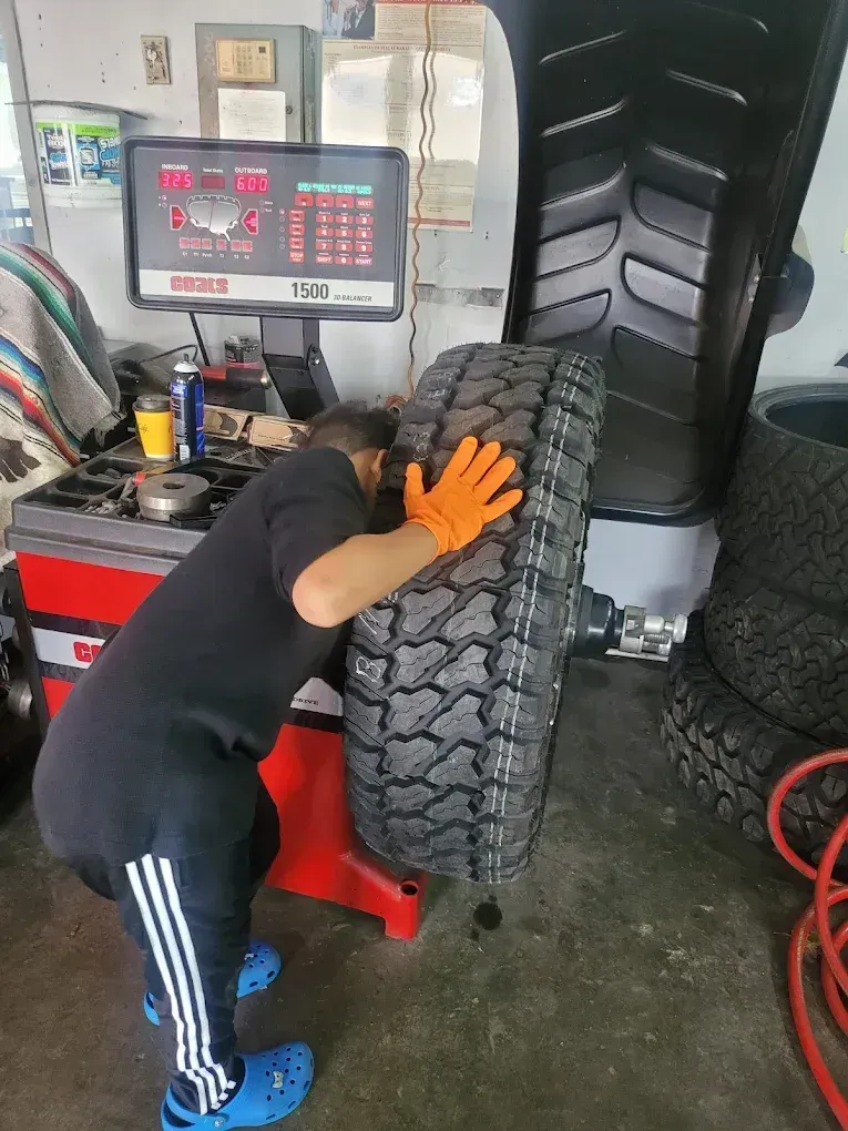 A person wearing orange gloves and blue clogs balances a large vehicle tire on a wheel balancing machine in a garage.