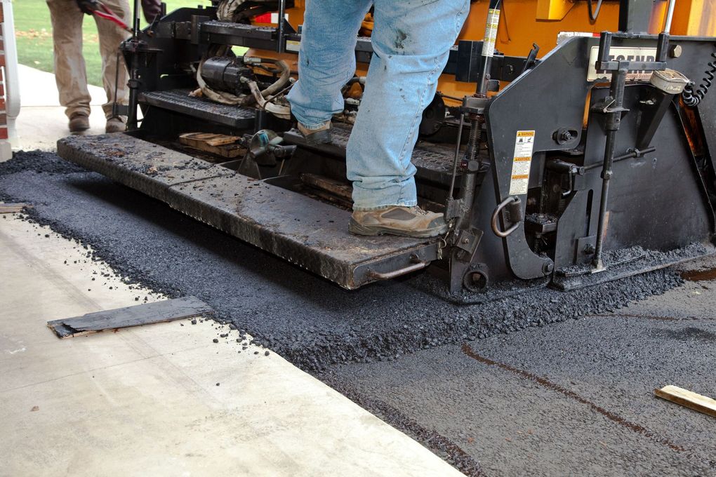 Asphalt paving machine laying black asphalt on a concrete surface. A person stands on the platform.