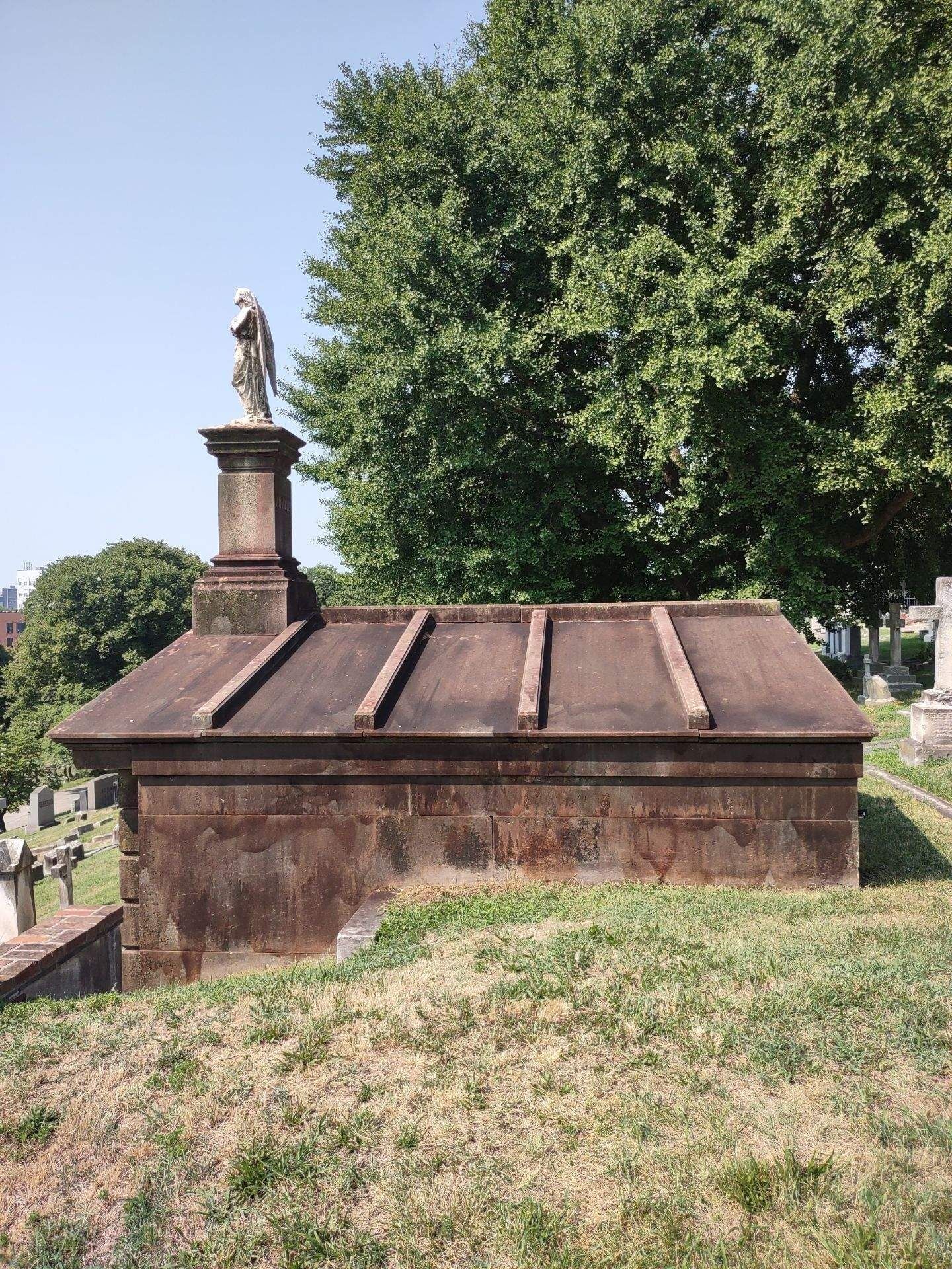 Stone mausoleum in a cemetery, with a statue on top. Sunny day.