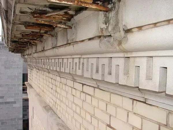 Close-up of a building's deteriorating exterior; white ceramic facade with brickwork and corroded metal beams visible.