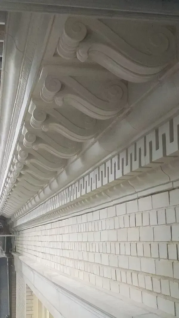 Ornate architectural detail: molded white cornice with Greek key pattern, above weathered brick wall.