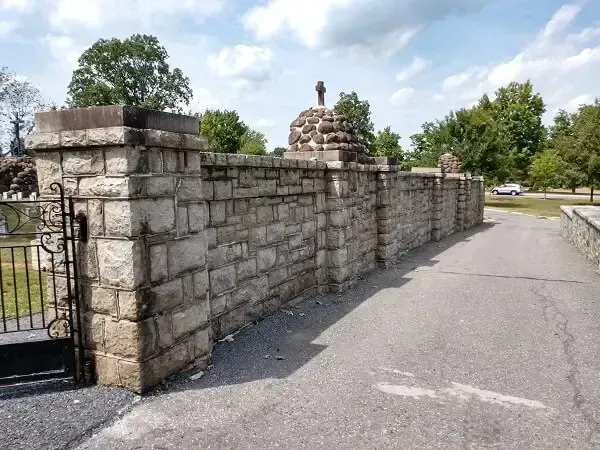 Stone wall surrounding cemetery entrance, road, and cloudy sky.