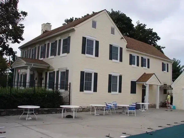 Two-story beige house with black shutters, several tables and chairs on a patio beside a pool.