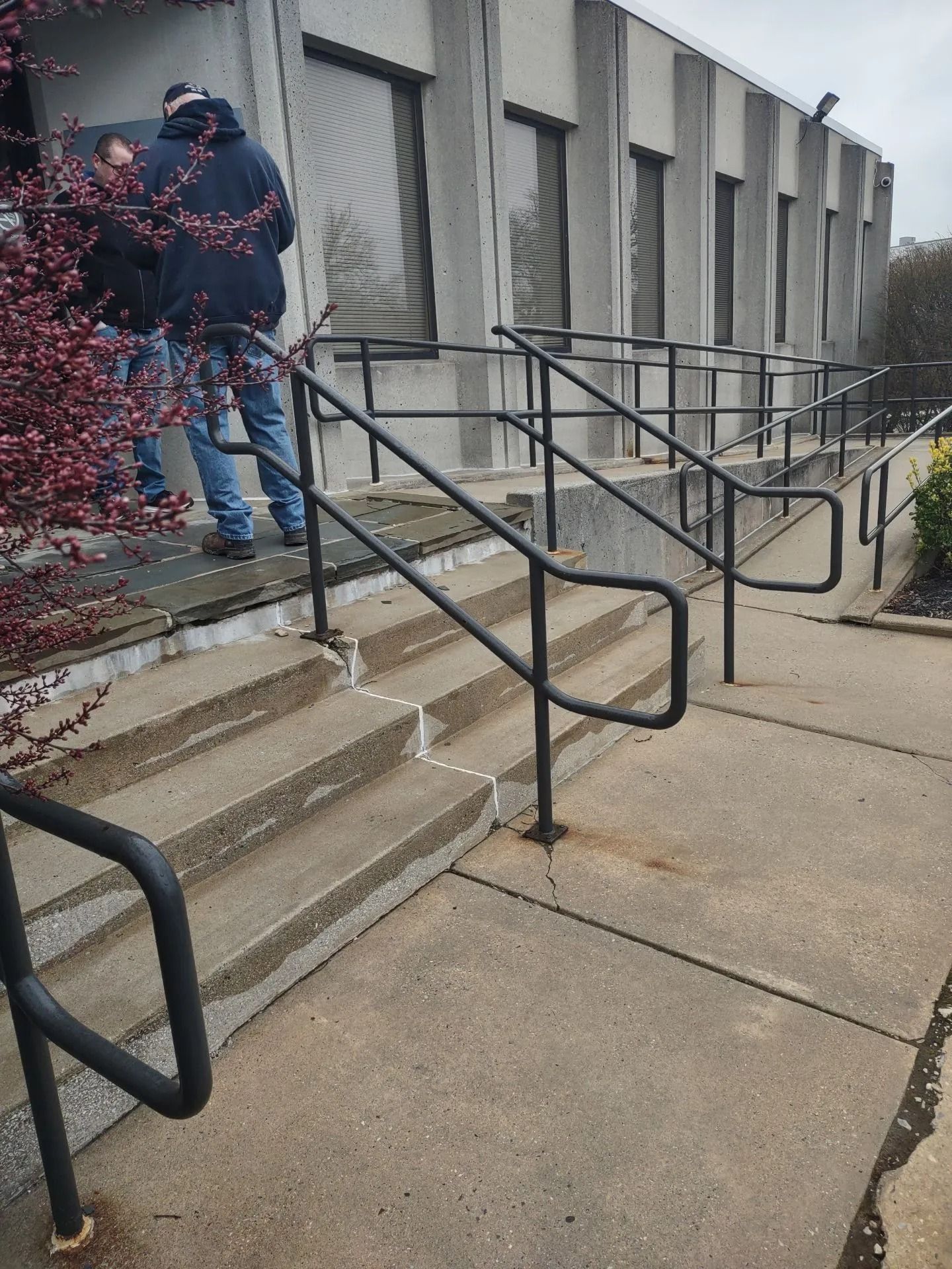 Concrete stairs with black handrails leading up to a building entrance. Two people stand near the door.