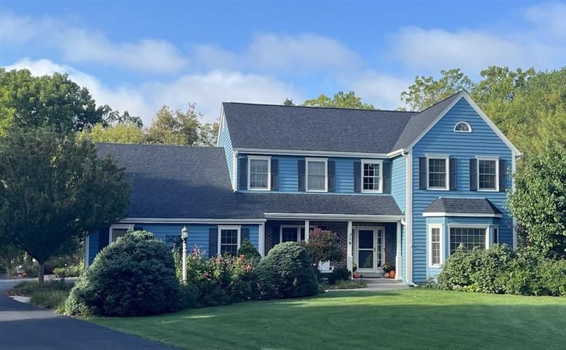 Blue two-story house with black shutters, green lawn, and trees under a partly cloudy sky.
