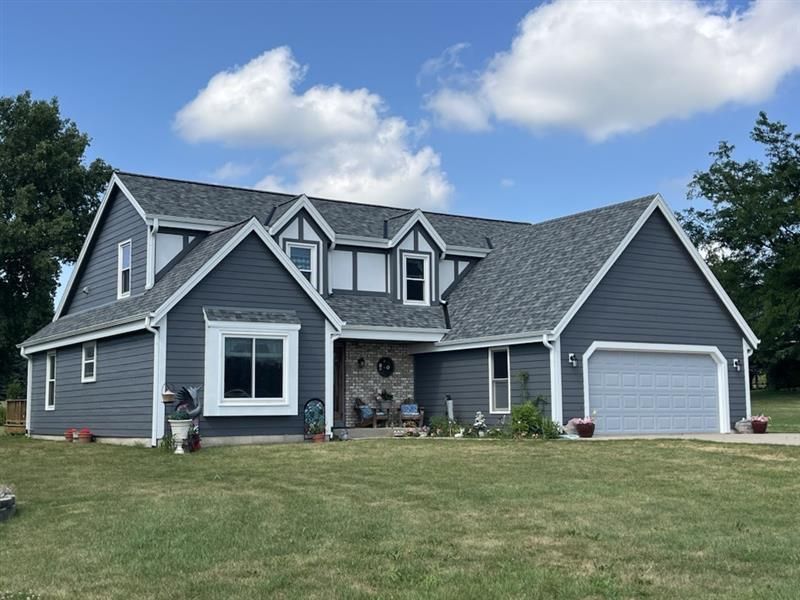 Blue house with white trim, gray roof, and a two-car garage on a green lawn under a blue sky.