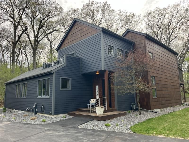Modern two-story house with dark blue and brown siding, surrounded by trees and gravel landscaping.