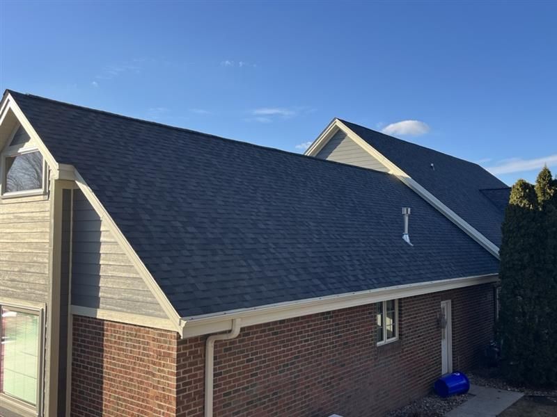 Dark gray shingled roof on a two-story brick and wood house against a blue sky.