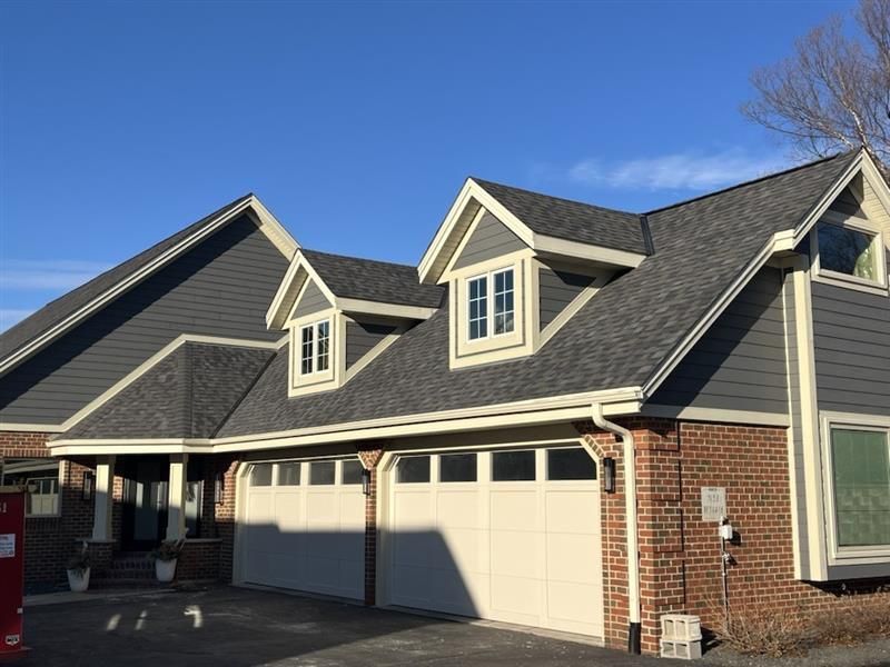 House with gray roof and siding, two dormers, red brick, and white garage doors.