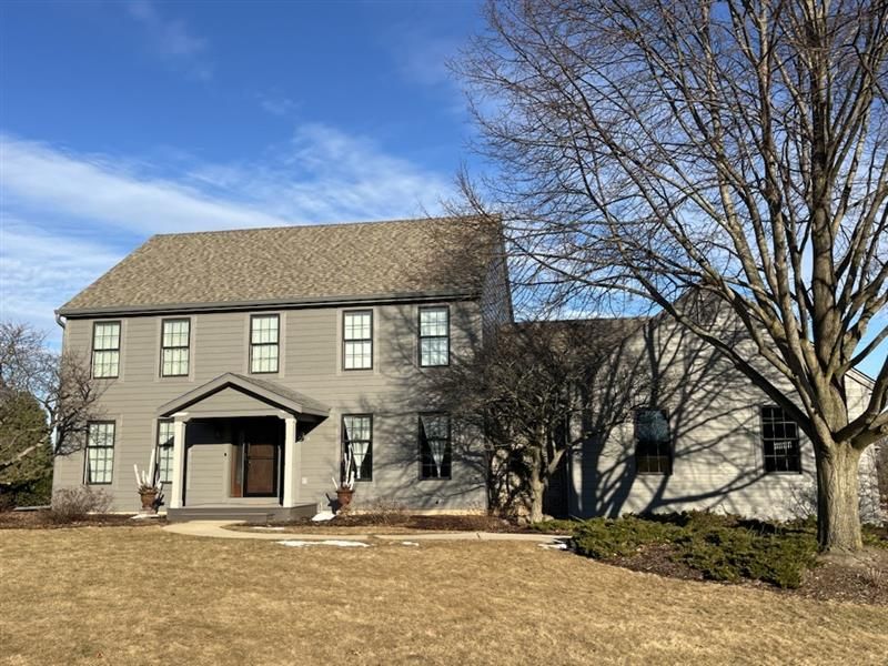 Two-story gray house with brown roof on a sunny day. Bare tree and brown grass in the foreground.