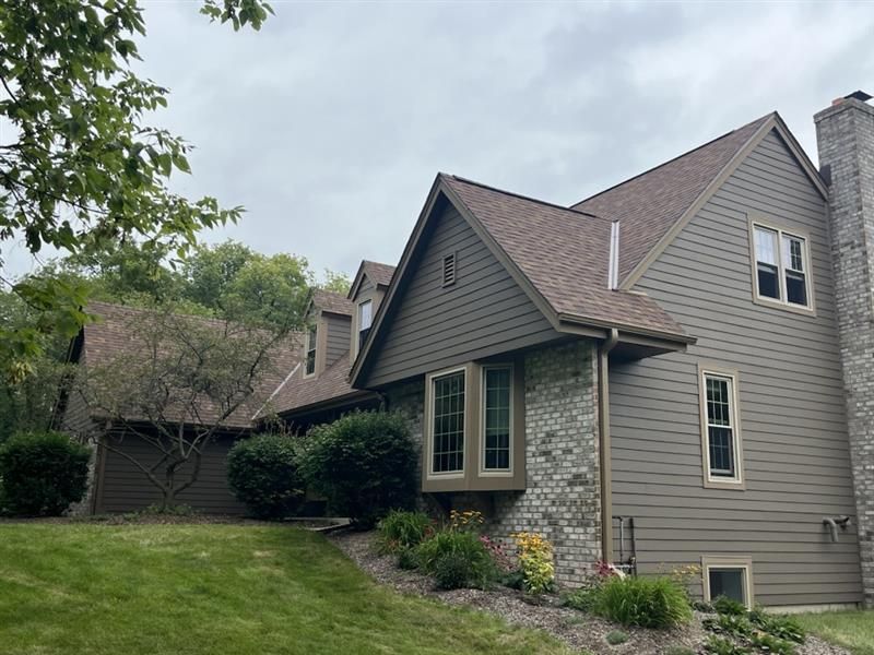 Two-story house with brown roof, green siding, brick accents, and chimney, on a grassy lawn under a cloudy sky.