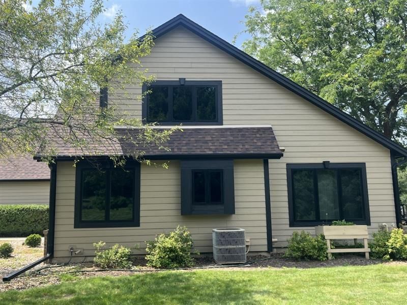 Beige house with black trim, windows, and roof against a blue sky, with green grass and shrubbery.