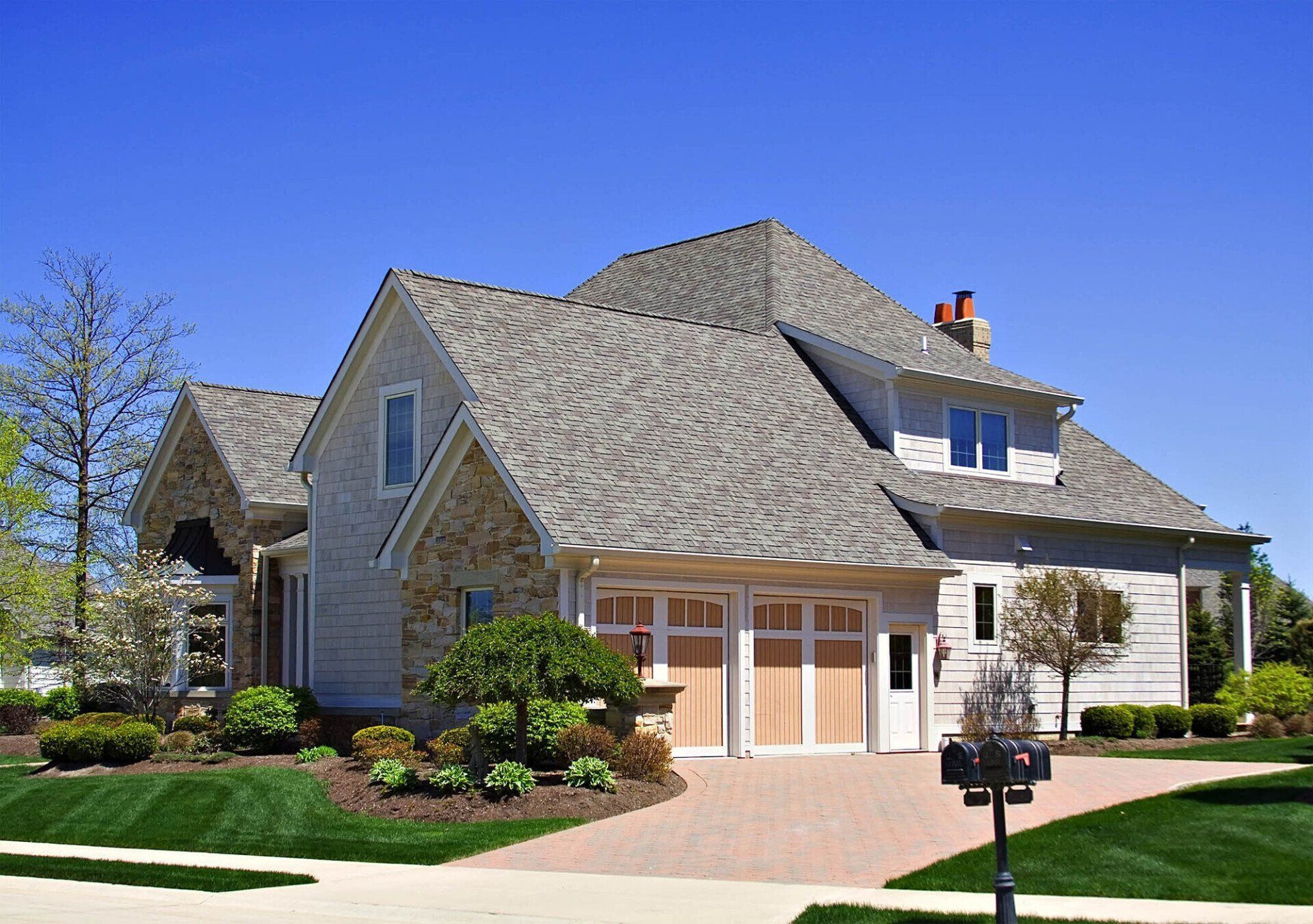 Two-story house with light siding, stone accents, and a red brick driveway against a blue sky.