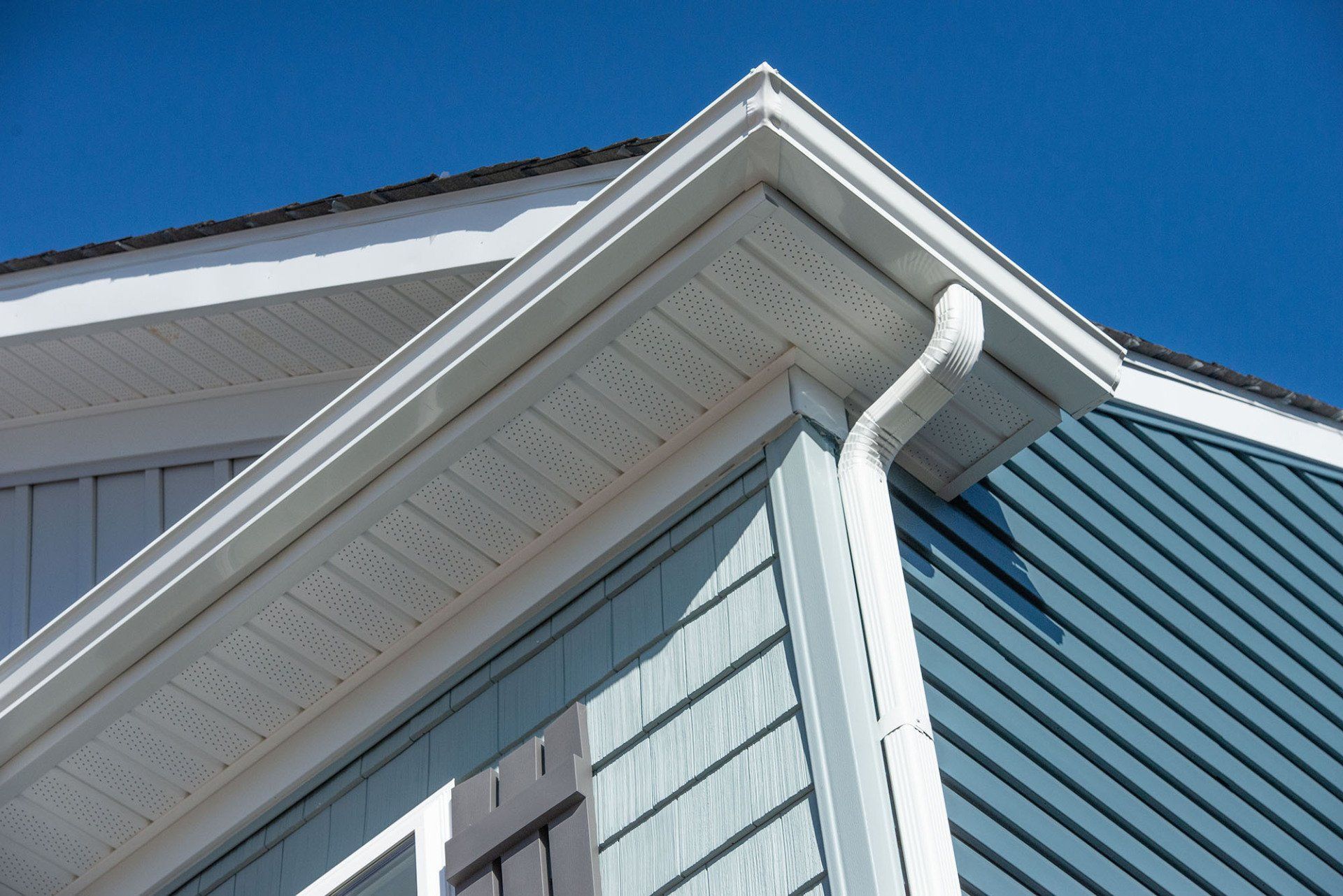 Corner of a house with blue siding, white trim, and a gutter against a clear blue sky.