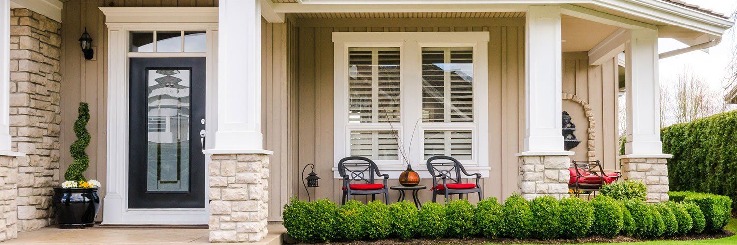 Front porch of a beige house with a black door, white columns, and a green hedge. Two chairs sit near a window.