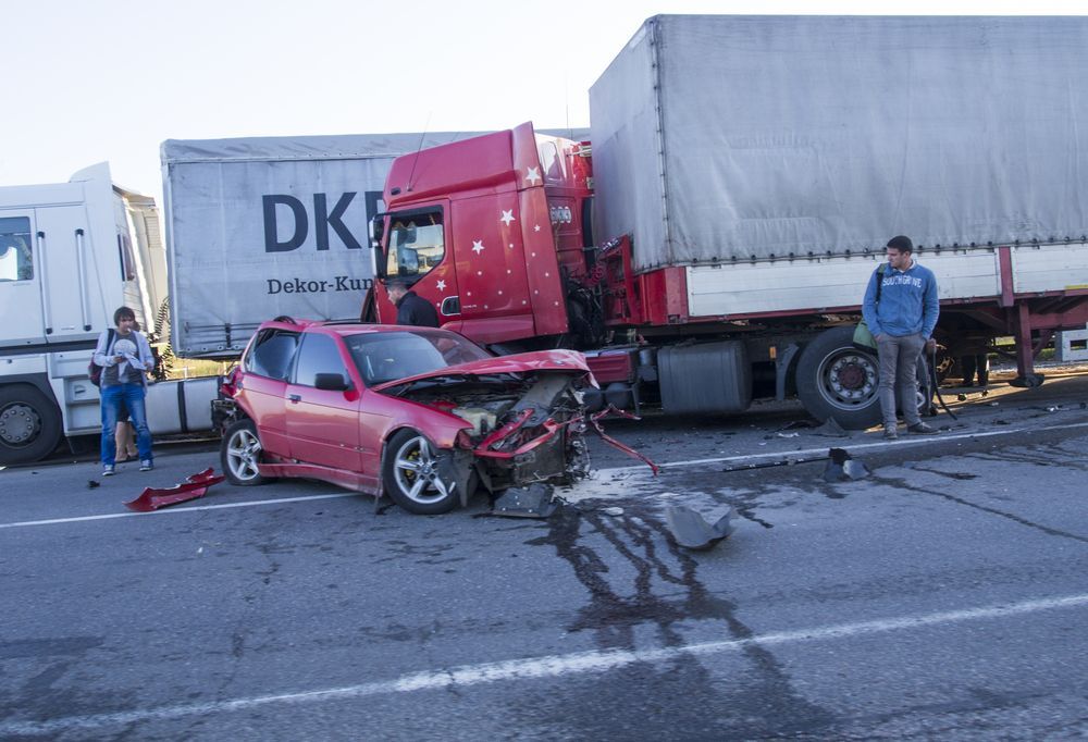 Red car heavily damaged after collision with a red semi-truck; two people stand nearby on the road.