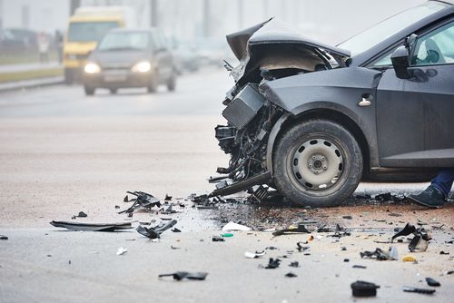 Damaged black car after a collision, debris on road, other vehicles in background.