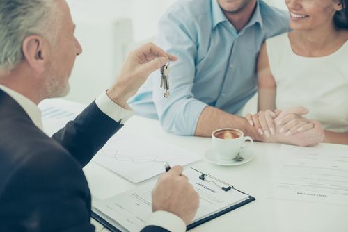 Real estate agent handing keys to a couple at a table; documents and coffee present.