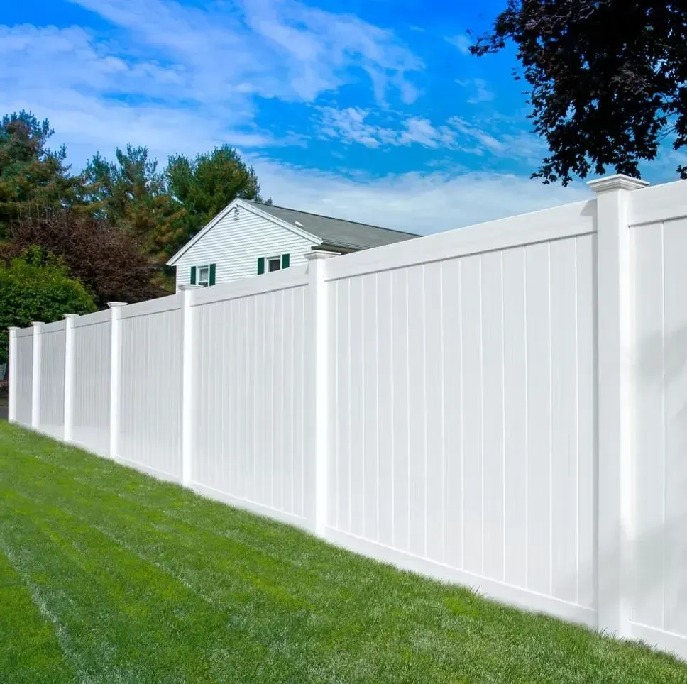 A long, white vinyl privacy fence stands in a grassy yard under a blue sky, with a house visible in the background.