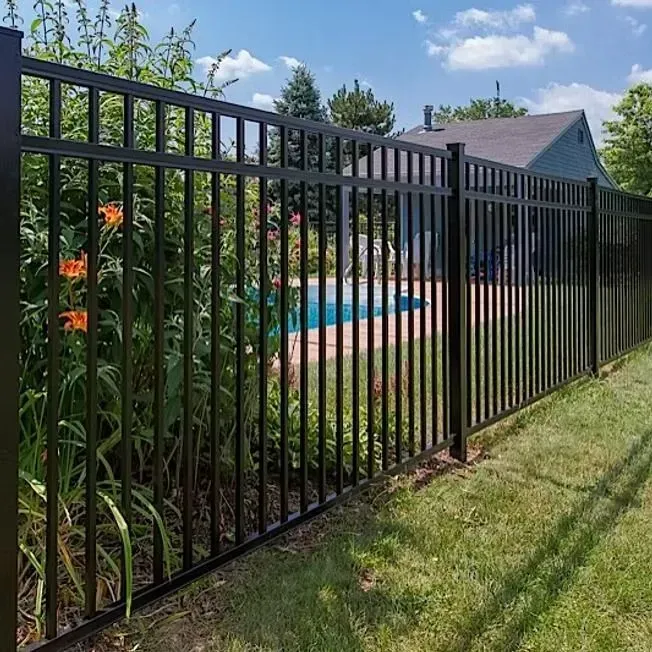A black metal fence encloses a green lawn and swimming pool, with orange flowers in the foreground under a blue sky.