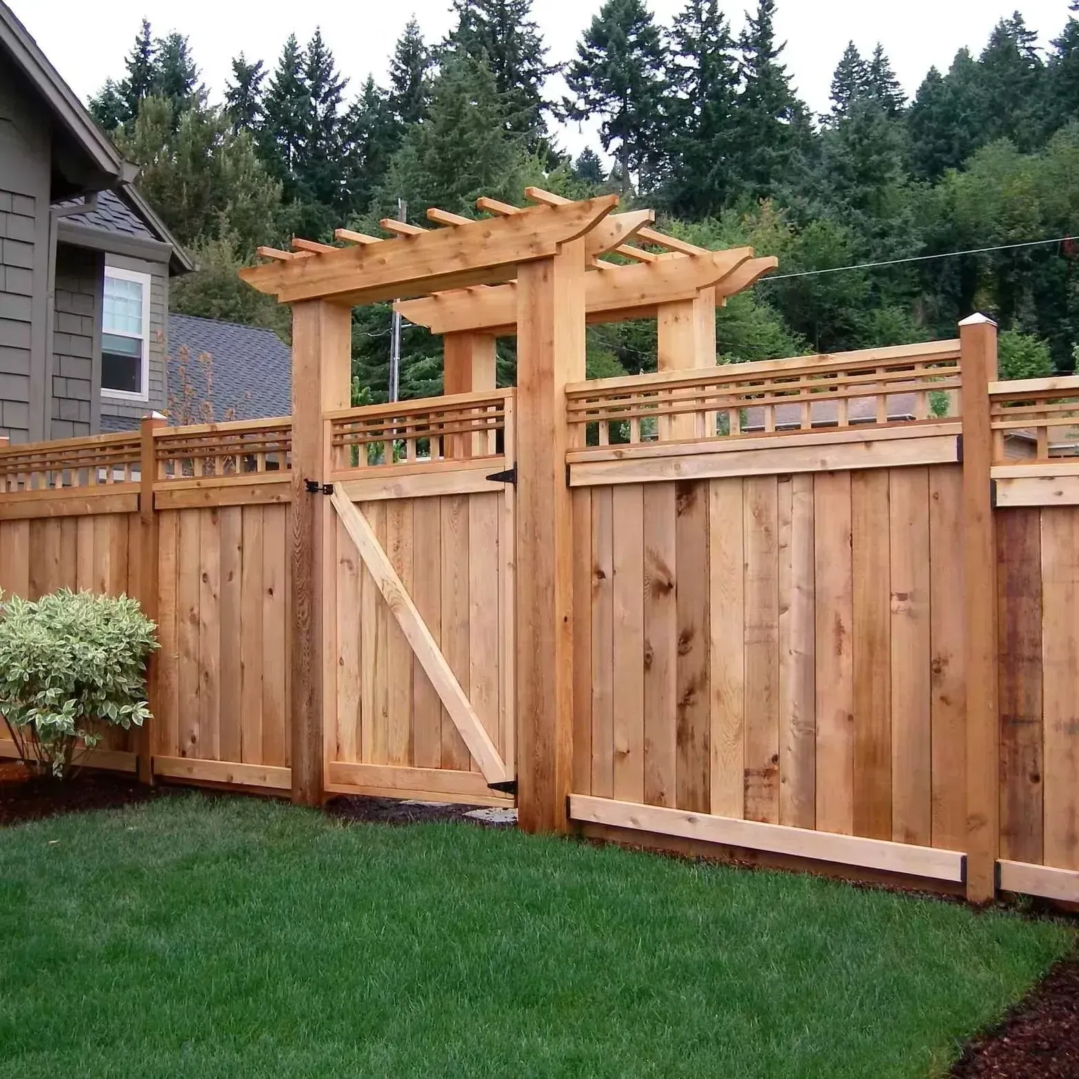 A wooden fence with a matching gate topped by a pergola, situated next to a house with green trees in the background.