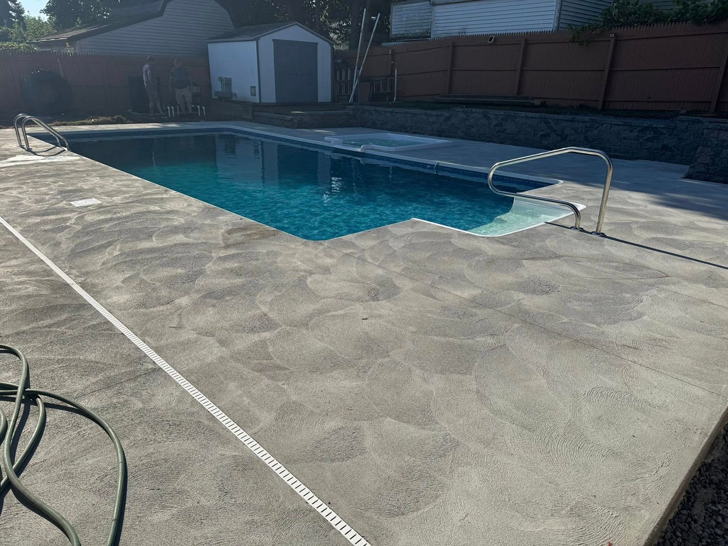 A rectangular backyard swimming pool with dark blue water and a concrete deck, bordered by a fence and a small shed.