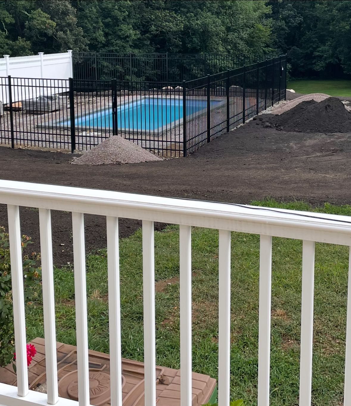 A swimming pool surrounded by a black metal fence sits in a backyard with piles of dirt and fresh landscaping in progress.