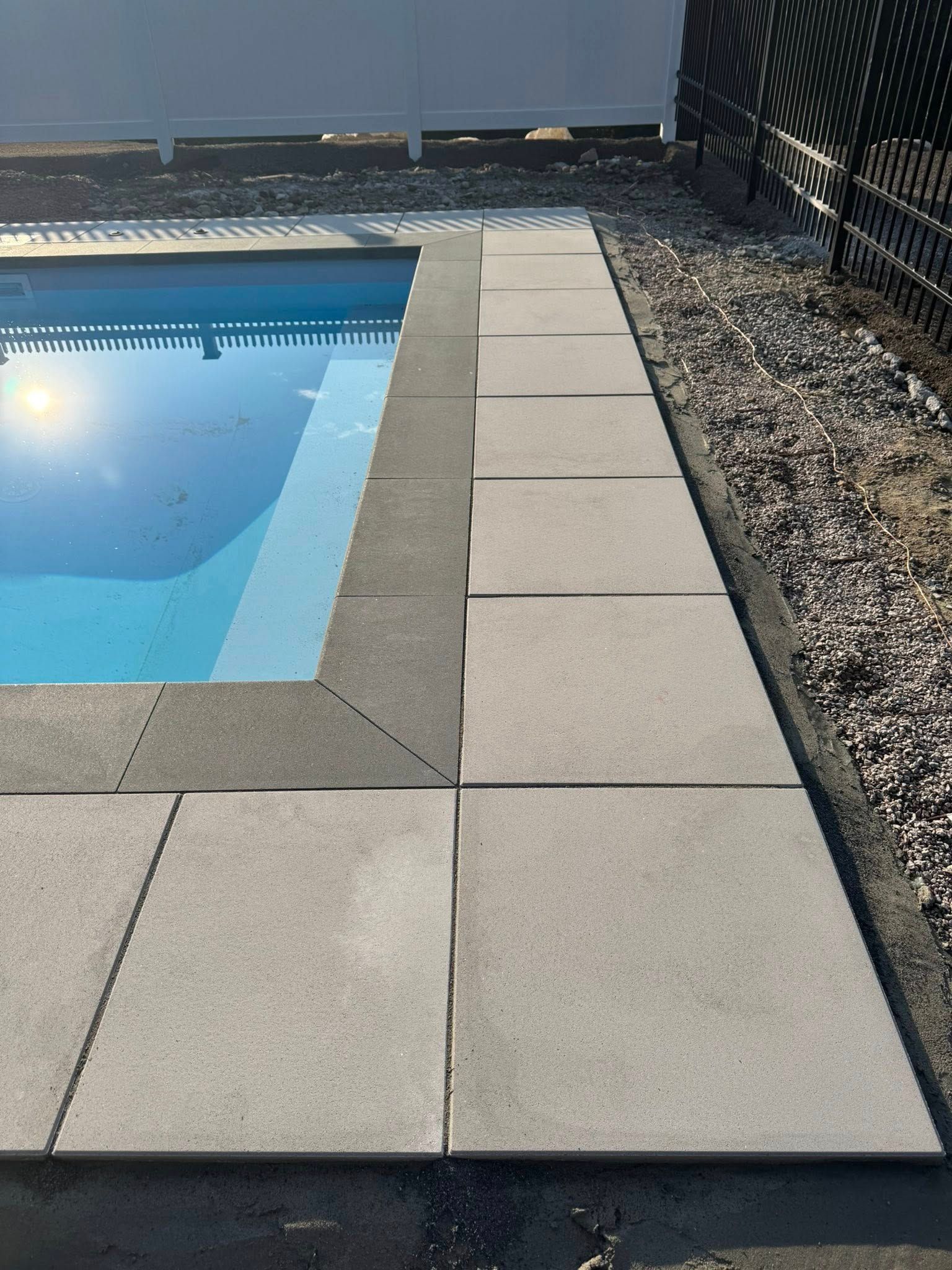 Light-colored concrete pavers surround a blue swimming pool, bordered by a gravel path and a black fence.