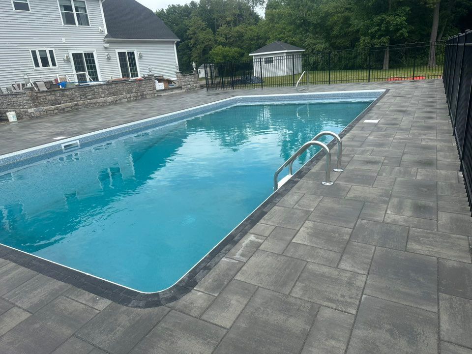 A rectangular swimming pool with blue water and metal ladders, surrounded by a grey stone patio and a black fence.