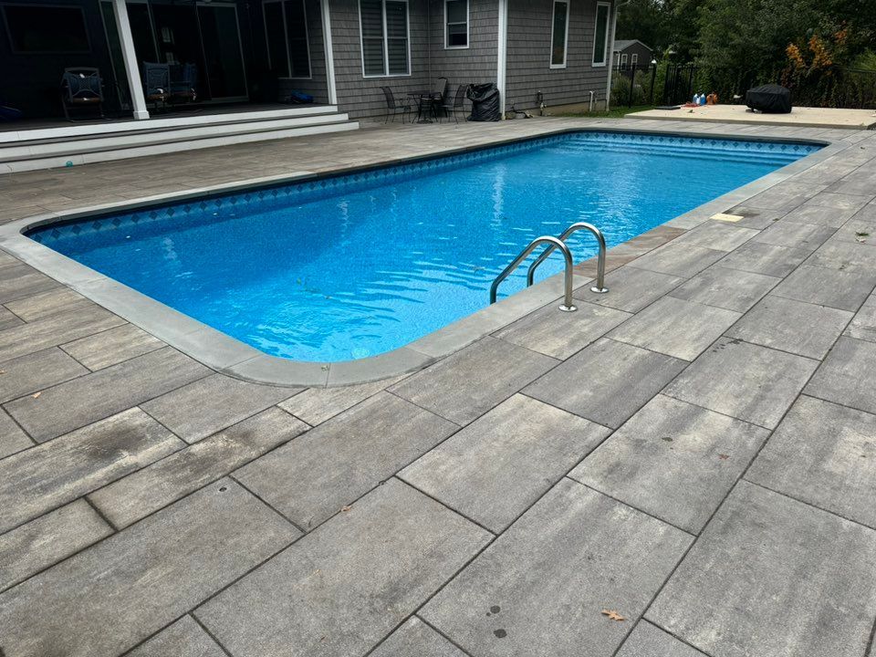 A rectangular swimming pool with bright blue water and a silver ladder, surrounded by light gray stone pavers.
