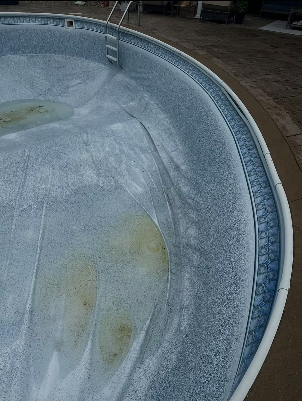 Empty, weathered in-ground swimming pool with stained liner and ladder, viewed from above.