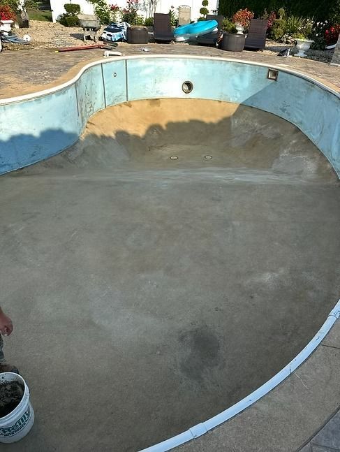 An empty, concrete swimming pool, painted blue on the sides, with a surrounding patio and landscaping in the background.
