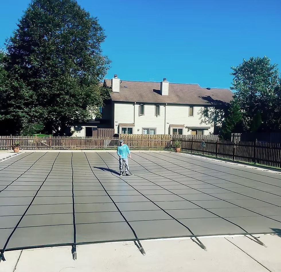 Person sweeping a large pool cover in front of a multi-unit building with a blue sky.