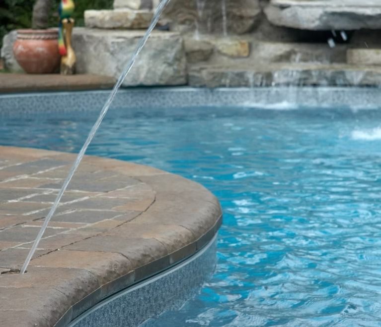Water fountain spraying into a blue swimming pool, with a stone border and waterfall in the background.