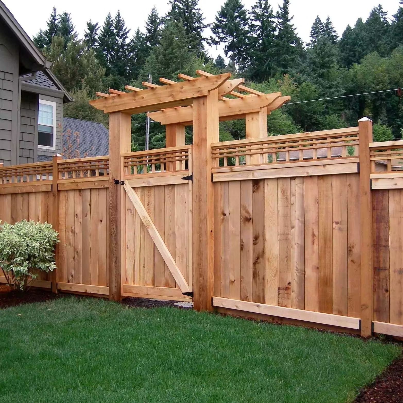 Wooden fence with gate, pergola above gate, green grass lawn, trees in background.