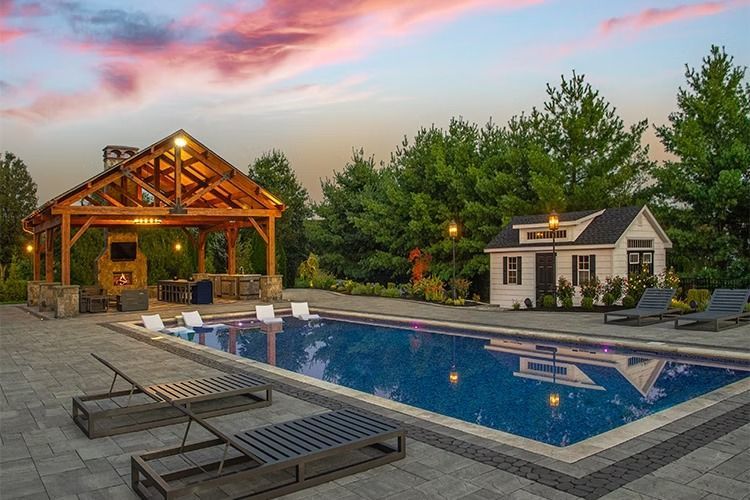 Pool with a wooden gazebo, a small white building, and lounge chairs at dusk under a colorful sky.