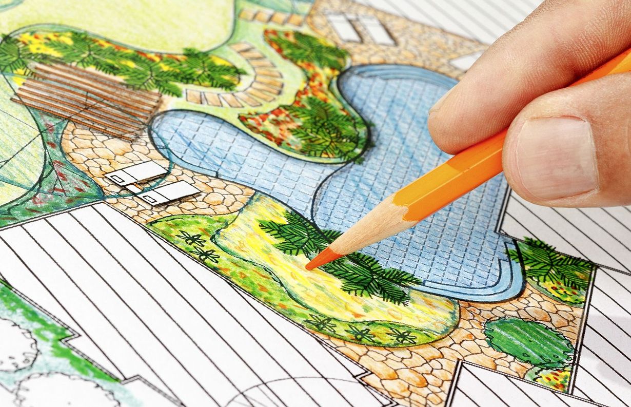 A hand uses an orange pencil to sketch a landscape design plan featuring a swimming pool and patio area.