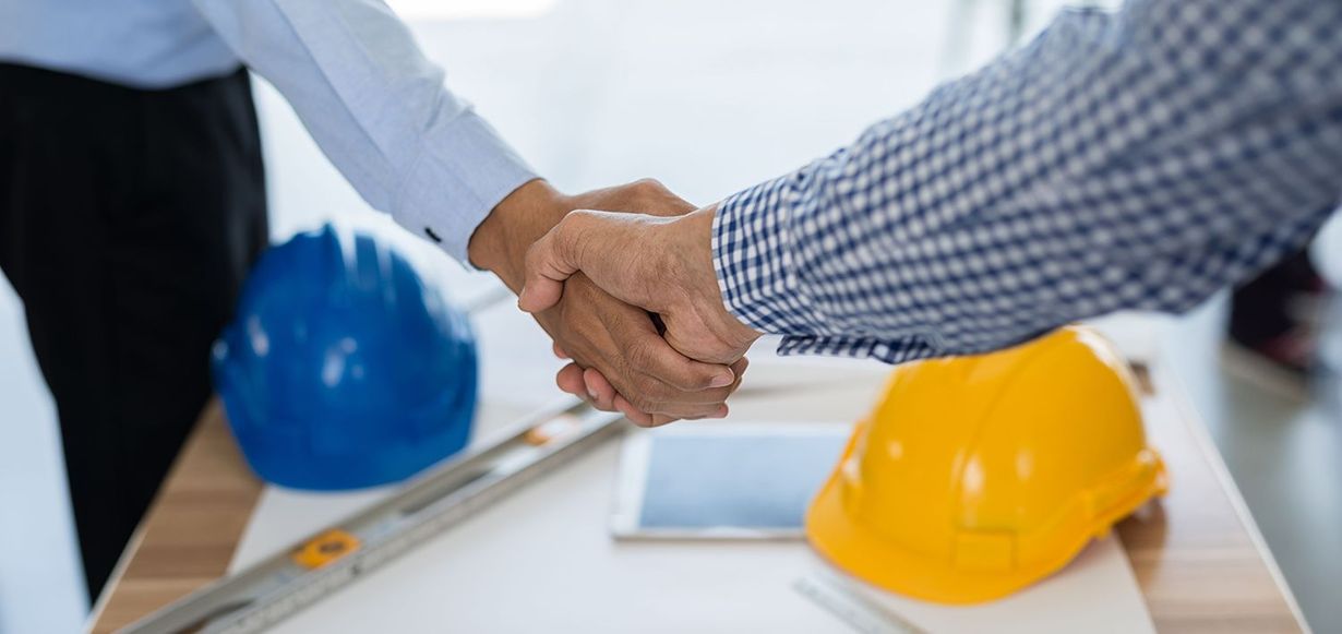 Two people shake hands over construction plans on a table with blue and yellow hard hats.