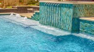 A turquoise-tiled spa wall with a waterfall flowing into a blue swimming pool with small water jets.