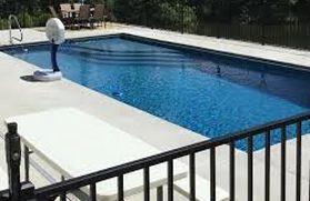 A rectangular blue swimming pool with a basketball hoop, white folding table, and black metal fence on a concrete deck.