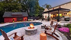 A backyard scene featuring a blue swimming pool, a circular stone fire pit with flames, and wooden chairs on a patio.
